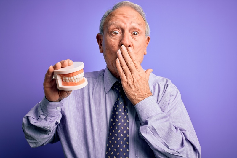  A gray-haired man holding dentures while covering his mouth in shock